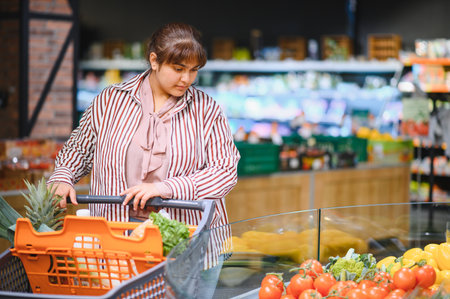 Young Indian customer pushing a shopping cart while selecting fresh, ripe tomatoes in a vibrant supermarket aisle filled with produceの写真素材