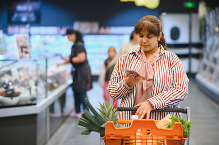 Young Indian woman consulting her smartphone while pushing a shopping cart full of fresh food in a supermarketの写真素材
