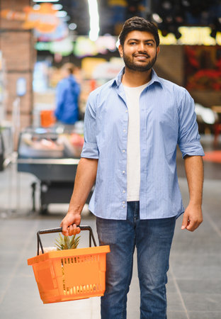 Young Indian man carrying shopping basket while walking through grocery store, smiling and looking awayの写真素材