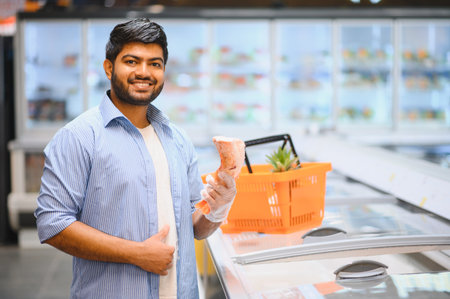 Young Indian man shopping for groceries, holding meat in a supermarket, smiling and giving thumbs upの写真素材