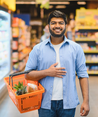 Happy indian man holding a shopping basket filled with fresh groceries, smiling brightly while enjoying his time in a bustling supermarketの写真素材
