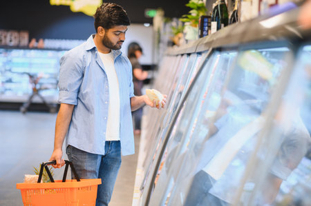 Young Indian man shopping for groceries, holding a basket and choosing frozen food from a refrigerated display case in a supermarketの写真素材