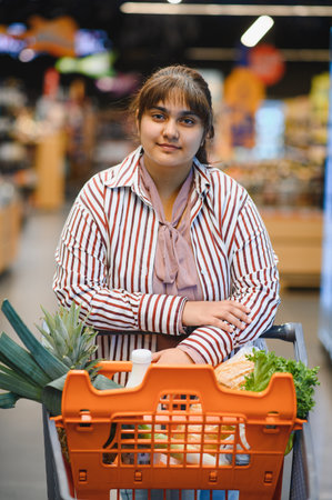 Young Indian woman pushing a shopping cart overflowing with fresh groceries while smiling in a vibrant supermarket aisleの写真素材