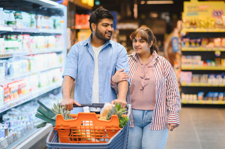 Young Indian couple pushing a shopping cart filled with groceries while enjoying their time together in a vibrant supermarket aisleの写真素材