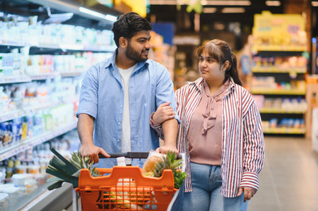 Young Indian couple pushing a shopping cart and chatting while selecting fresh groceries in a bright, organized supermarket aisleの写真素材