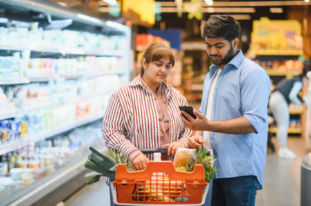 Indian couple consulting a smartphone while selecting fresh groceries in a supermarket, enjoying modern shopping technology togetherの写真素材