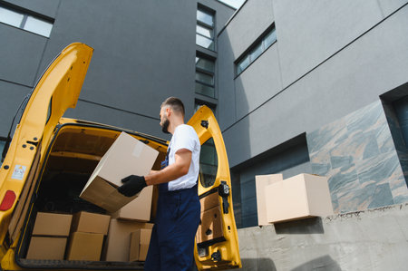 Courier gloves wearing loading cardboard boxes into a yellow delivery van parked near a modern...