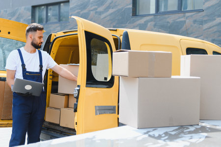 Courier wearing overalls, checking packages on a laptop while loading a bright yellow delivery van...