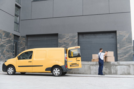 Courier arranging cardboard boxes near a yellow delivery van parked in front of a modern building, preparing for shipping and handlingの写真素材