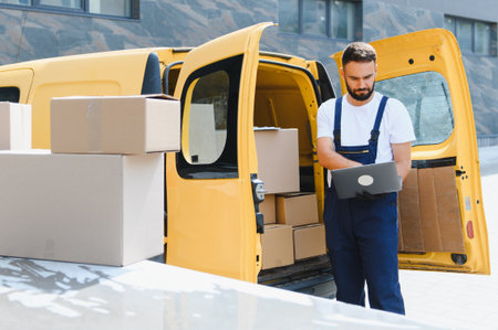 Delivery worker using laptop next to yellow delivery van loaded with cardboard boxes, managing...