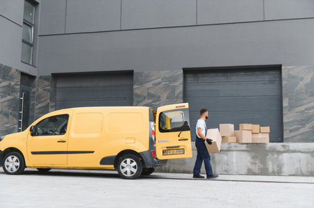 Courier wearing protective face mask carrying cardboard box from a yellow delivery van parked next to a warehouseの写真素材