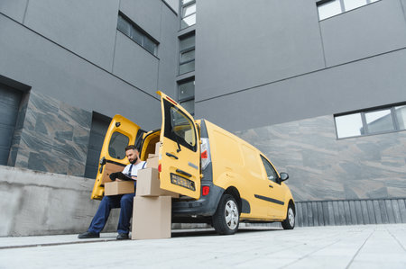 Delivery man checking delivery notes sitting near his yellow van full of cardboard boxes with open...