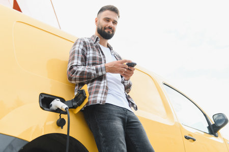 Delivery man using a smartphone while charging his electric van at a charging station, embracing sustainable and eco friendly transportationの写真素材
