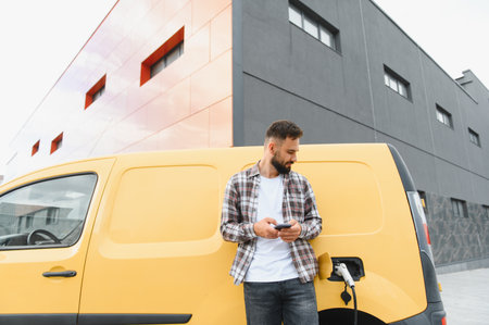 Delivery driver using a smartphone while charging an electric van outside a modern building, showcasing sustainable transportation technologyの写真素材