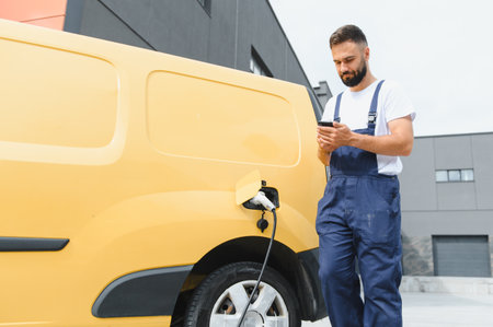 Courier checking his smartphone while charging an electric delivery van, showcasing modern technology and sustainable transportation practicesの写真素材