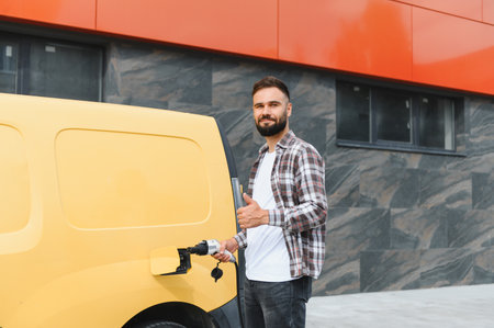 Delivery driver charging an electric van and giving a thumbs up in front of a modern building, showing a commitment to sustainable transportationの写真素材