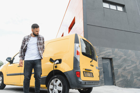 Delivery driver waiting beside a yellow electric van, charging in front of a sleek modern building...