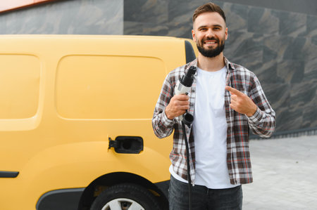 Delivery driver holding charging cable and pointing at his electric delivery van, promoting sustainable transportationの写真素材
