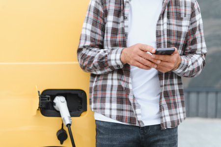 Driver using mobile phone while charging electric car at charging station, focus on sustainable transportation and technologyの写真素材