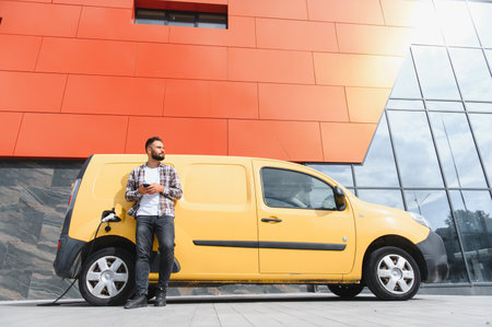 Delivery driver using smartphone while his electric van is charging in front of modern building