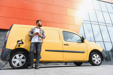 Delivery driver using smartphone while his electric van is charging in front of modern buildingの写真素材
