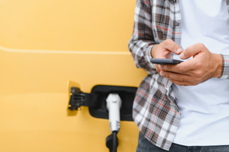 Truck driver using a smartphone while his electric vehicle is charging, representing the future of transportation and logisticsの写真素材