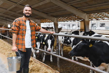Young farmer holding a metal bucket while observing cows in a modern dairy barn, ensuring care for livestock and milk productionの写真素材