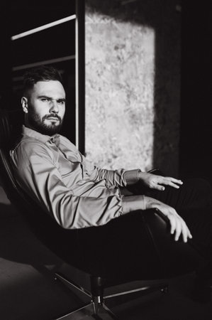 Serious bearded man wearing a shirt, sitting on a black office chair, intense look, dramatic lighting, black and white effectの写真素材