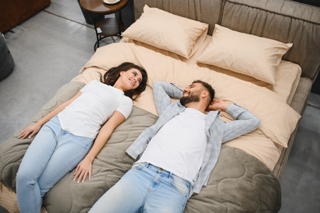 Happy young couple lying on a bed, smiling and looking at each other, enjoying a moment while choosing furniture for homeの写真素材