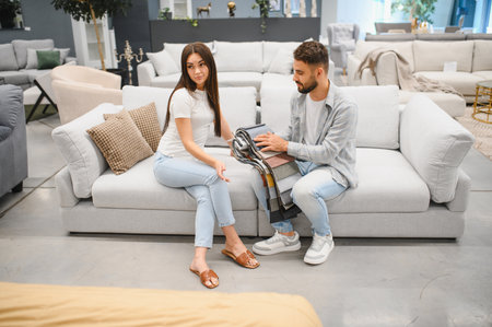 Young couple seated on a sofa in a furniture showroom comparing fabric swatches and discussing upholstery choices for their new homeの写真素材