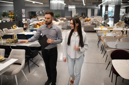 Male sales consultant with clipboard guiding a female client through a furniture store, pointing out dining room optionsの写真素材