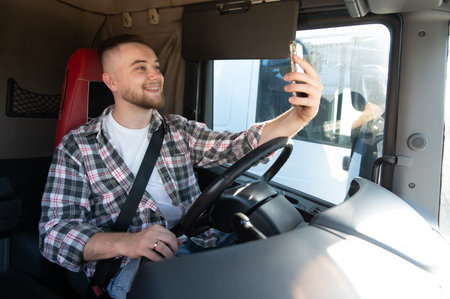 Happy young male truck driver smiling, wearing plaid shirt and seatbelt, taking selfie with...