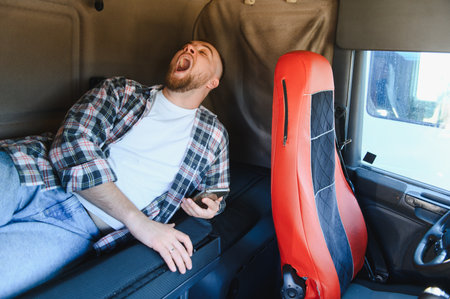 Truck driver feeling weary, yawning widely, taking a rest break on the sleeper berth inside the cabin of his heavy vehicleの写真素材