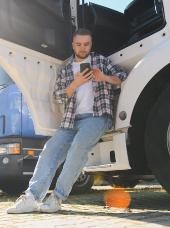 Young truck driver sitting on his truck step during a break, confidently using smartphone to check routes and messages on a sunny dayの写真素材