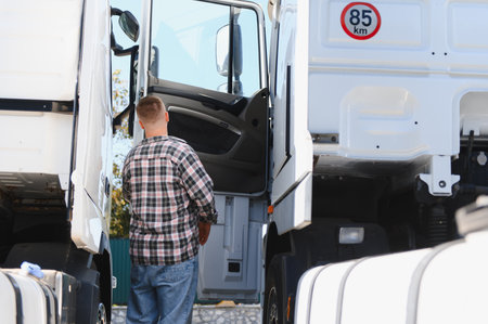 Truck driver in plaid shirt standing by open semi truck door, prepping for a long haul delivery on a daytime highway journeyの写真素材