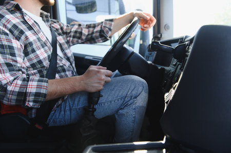 Truck driver wearing a plaid shirt and seatbelt, navigating a vehicle from inside the cabin, engaging safety measures for logisticsの写真素材