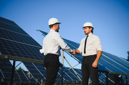Two happy bearded workers in uniform talking and shaking hands near solar panel farm.の写真素材