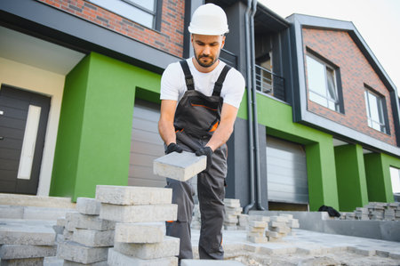 The master in gloves lays paving stones in layers. Garden brick pathway paving by professional paver worker. Laying gray concrete paving slabs in house courtyard on sand foundation base.の写真素材