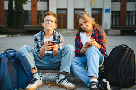 Two school little students small boy and girl sitting on stairs near modern school building. Pretty friends searching internet on smartphone and showing to friendの写真素材