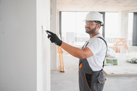 Male builder in work overalls plastering a wall using a construction trowel.