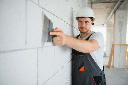 Worker in uniform plastering wall with putty knife indoors.