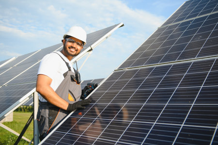 Portrait of Young Indian man technician wearing white hard hat standing near solar panels against...