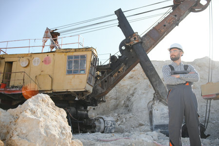 A worker in a helmet stands on the background of an excavator in a quarry.