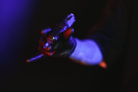 Closeup of tattooer master's hand in black glove holding machine for making tattoo art on body isolated on dark background in neon. Concept of art, job, hobby, professional equipment. Flyer for adの写真素材