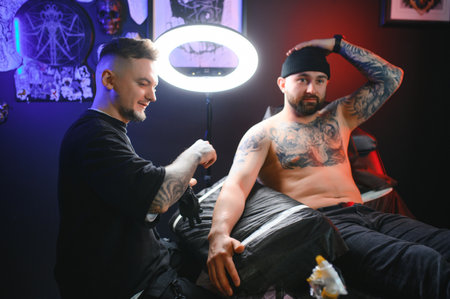 bearded tattoo artist working at his studio tattooing sleeve on the arm of his male client. Man getting tattooed by professional tattooist.の写真素材