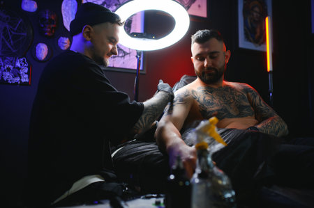Male tattoo artist holding a tattoo gun, showing a process of making tattoos on a male tattooed...
