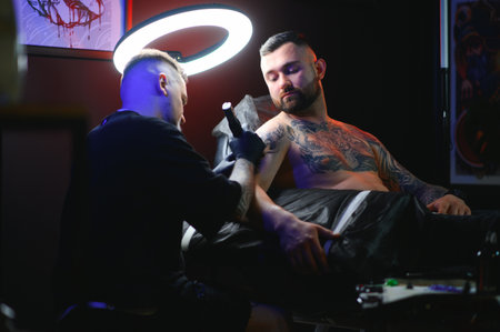 bearded tattoo artist working at his studio tattooing sleeve on the arm of his male client. Man getting tattooed by professional tattooist.の写真素材