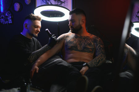 bearded tattoo artist working at his studio tattooing sleeve on the arm of his male client. Man getting tattooed by professional tattooist.の写真素材