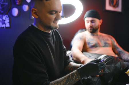 bearded tattoo artist working at his studio tattooing sleeve on the arm of his male client. Man...