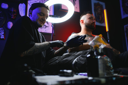 bearded tattoo artist working at his studio tattooing sleeve on the arm of his male client. Man getting tattooed by professional tattooist.の写真素材